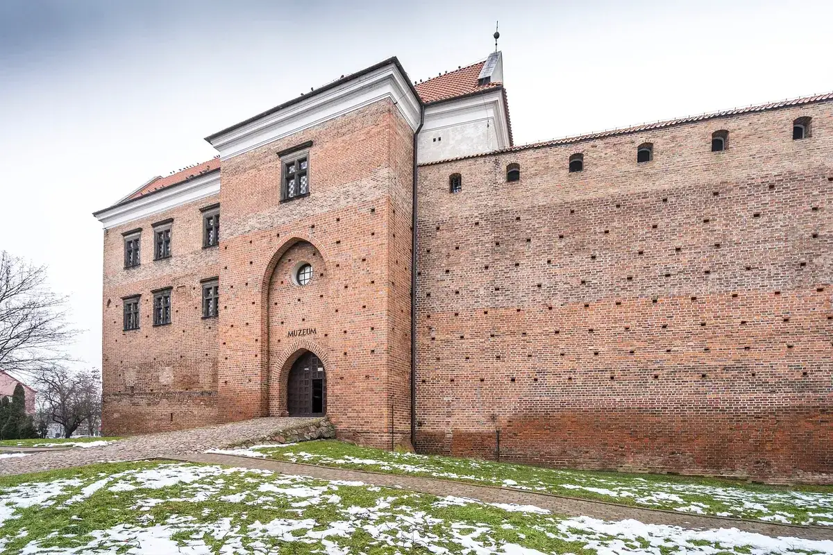 Zamek w Łęczycy, ceglana twierdza z wejściem do muzeum, otoczona zimową zielenią i resztkami śniegu.