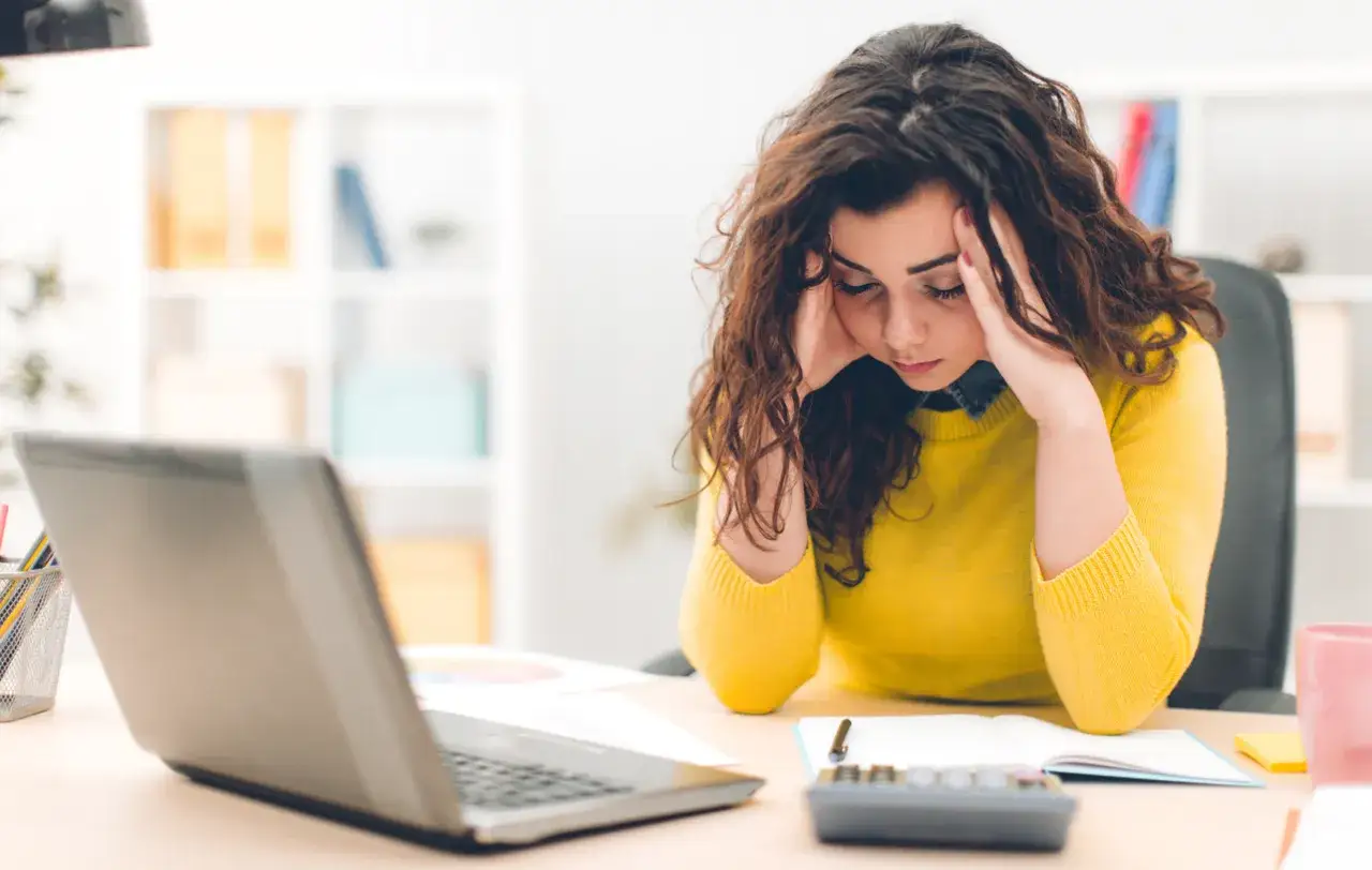 Donna con mal di testa e occhi stanchi, appoggiata alla scrivania con un laptop, quaderno e calcolatrice.