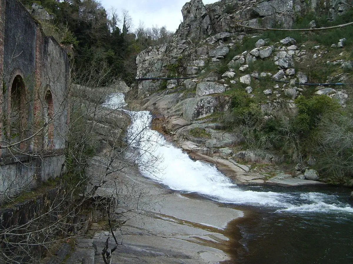 Cascada de auga branca na fervenza de Segade, con edificación antiga de ladrillo á esquerda e rochas e vexetación á dereita.