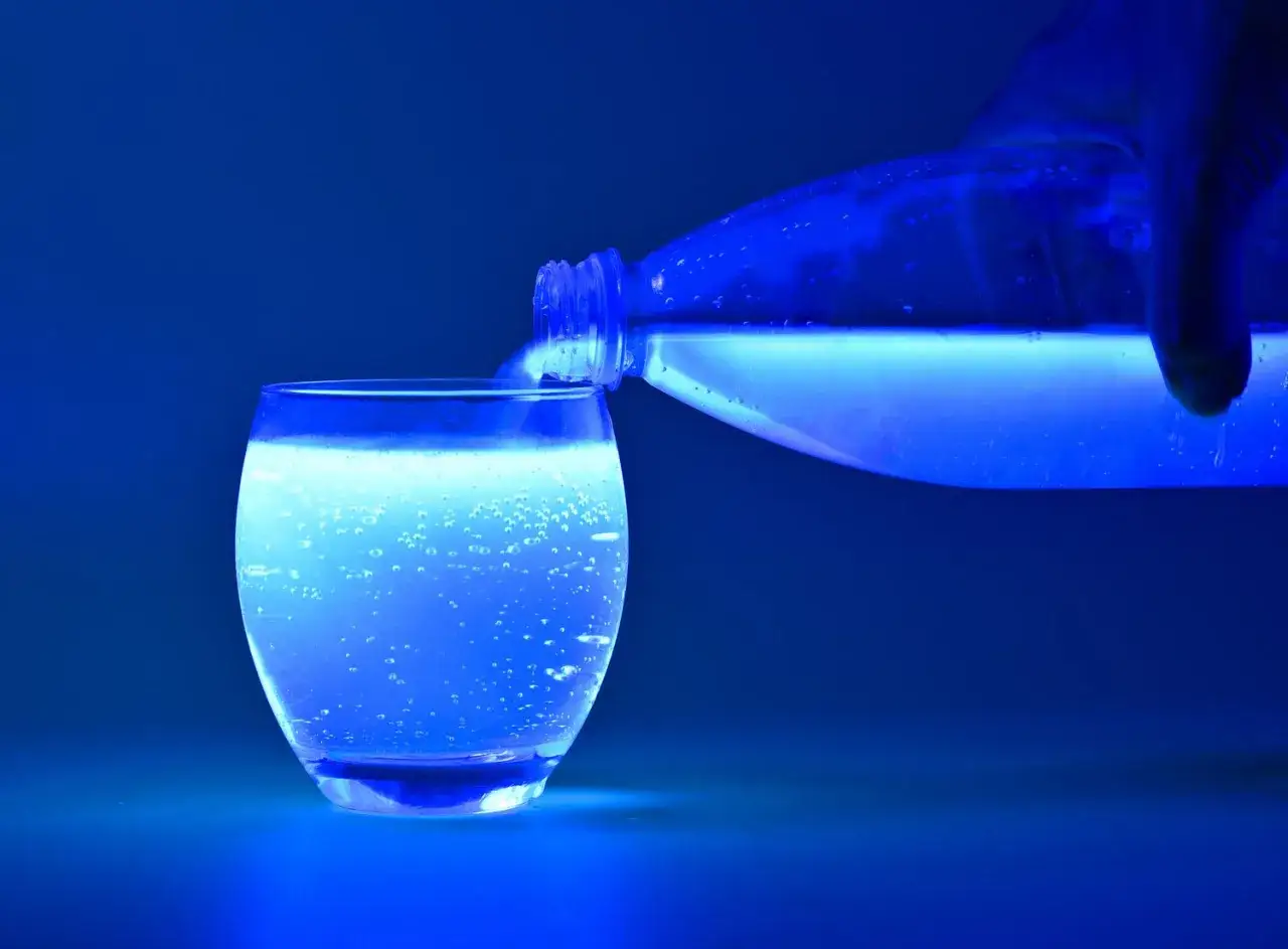 Ein Glas wird mit leuchtendem Tonic Water aufgef&uuml;llt. Die Fl&uuml;ssigkeit im Glas und in der Flasche schimmert intensiv blau.