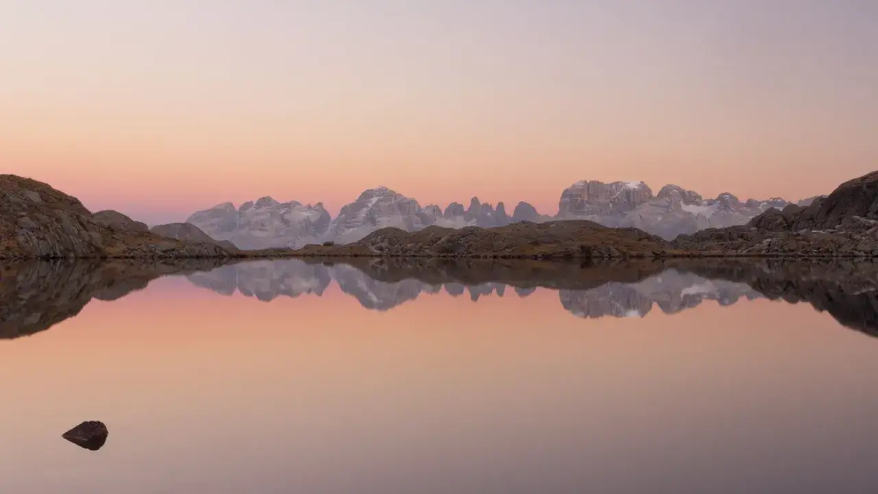 Alba sulle Dolomiti, un lago alpino riflette le cime innevate e il cielo rosa. Un sasso emerge dall'acqua, un punto di **max rive** in questo paesaggio sereno.