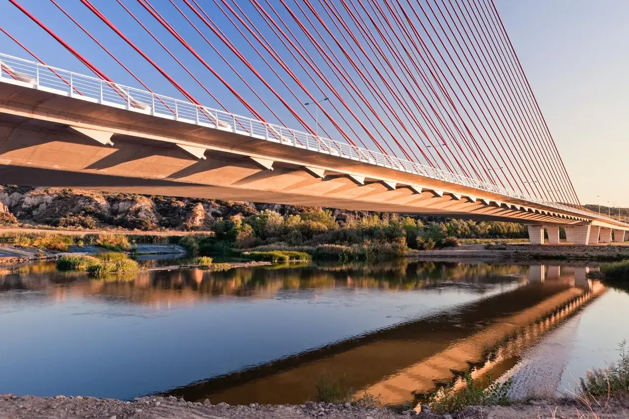 El puente de Castilla-La Mancha se refleja en el río, con sus cables rojos y el cielo azul.
