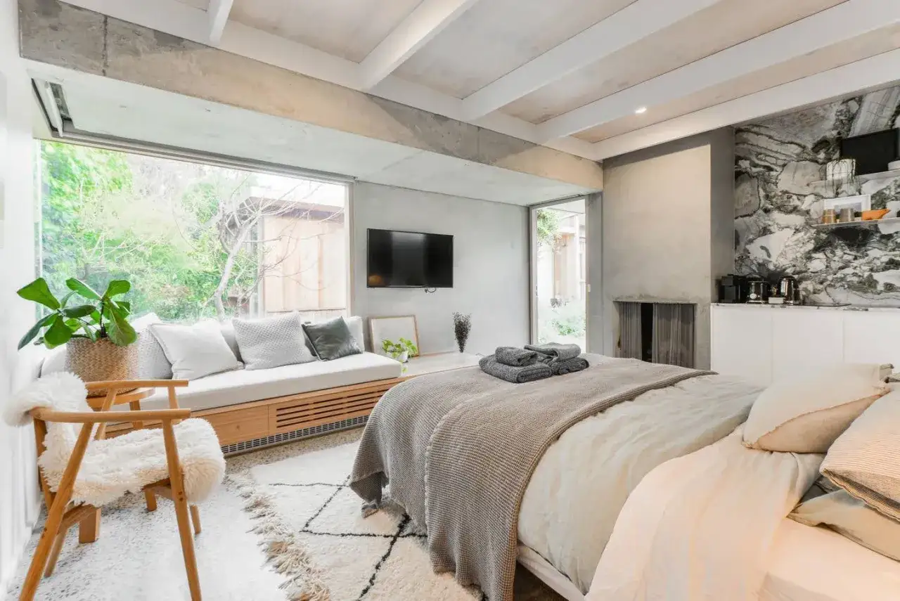Modernes Schlafzimmer mit Betonoptik an W&auml;nden und Decke. Gro&szlig;es Fenster, bequemes Bett, gem&uuml;tliche Sitzecke und ein Hauch von Luxus.
