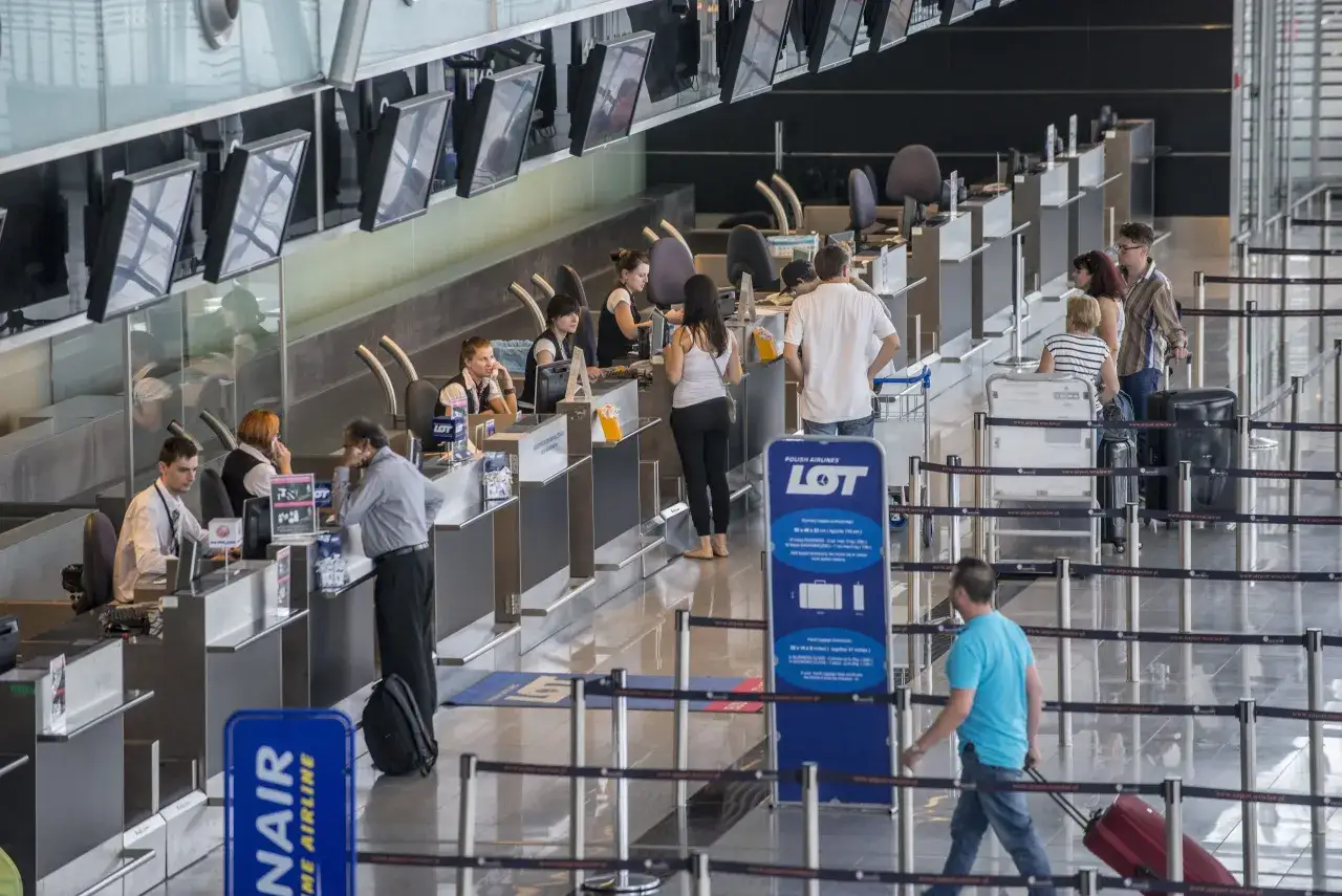 Kolejka do odprawy na lotnisku. W tle widać lady z pracownikami i pasażerami, a także ekrany informacyjne. Znak Ryanair w słońcu.