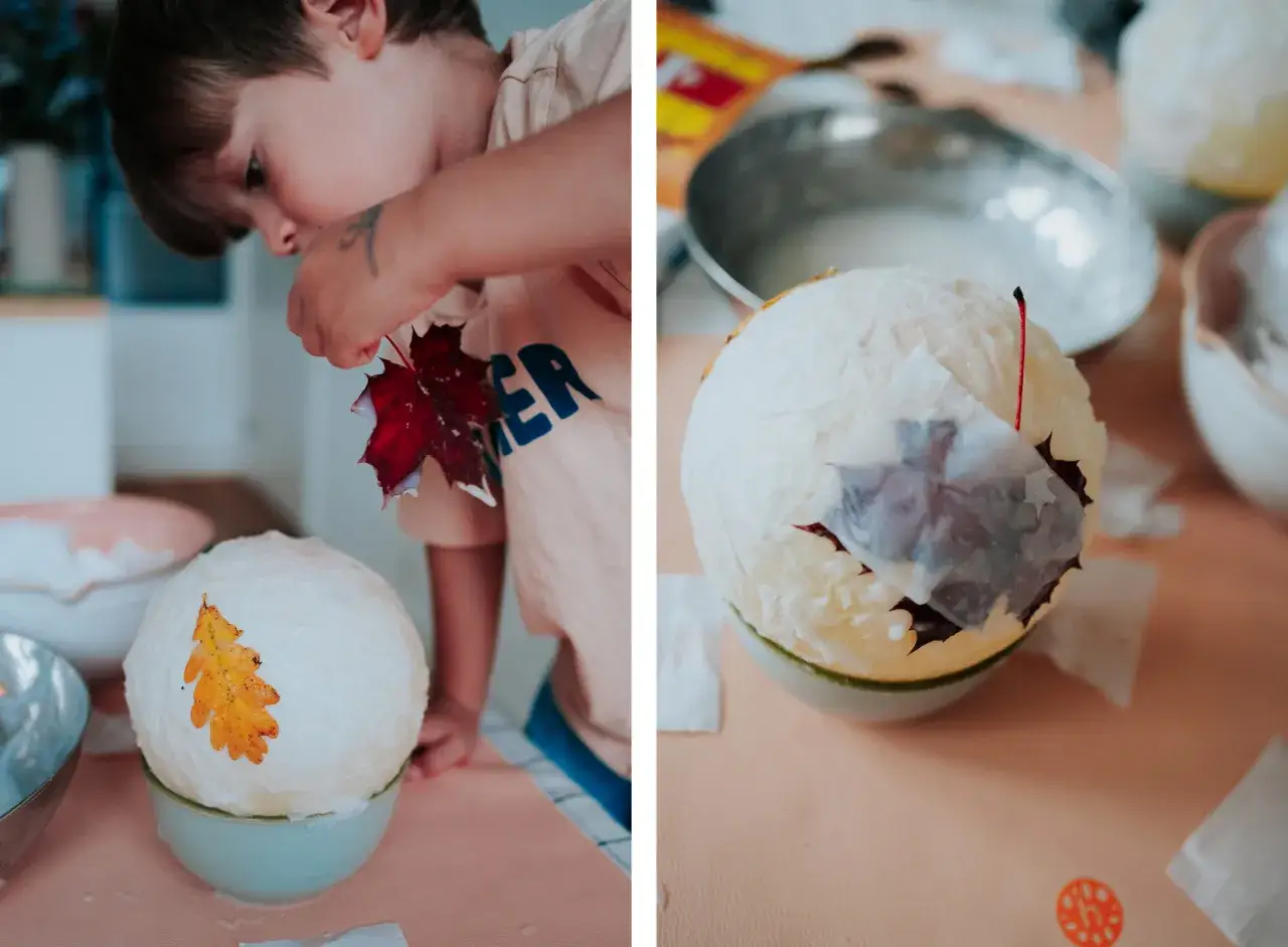 Kind bastelt mit Blättern und Papier einen Ballon. Ein gelbes Blatt ziert den weißen Ballon, ein rotes Blatt wird gerade angebracht.
