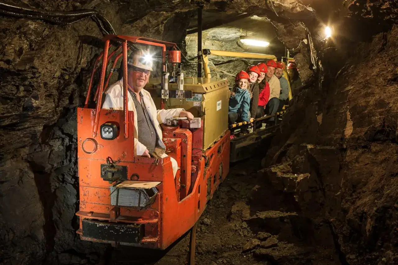 Eine Gruppe von Kindern und Erwachsenen f&auml;hrt mit einer Lore durch einen dunklen Stollen, um ein Bergwerk zu besichtigen.