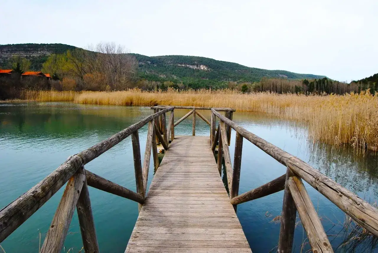 Muelle de madera que se adentra en la serena laguna de Uña, rodeada de juncos dorados y colinas verdes.
