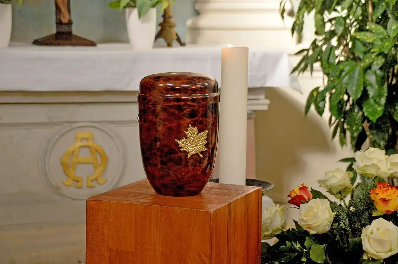 Elegancka urna z liściem klonu, symbolizująca prochy po kremacji, stoi obok świecy i kwiatów.