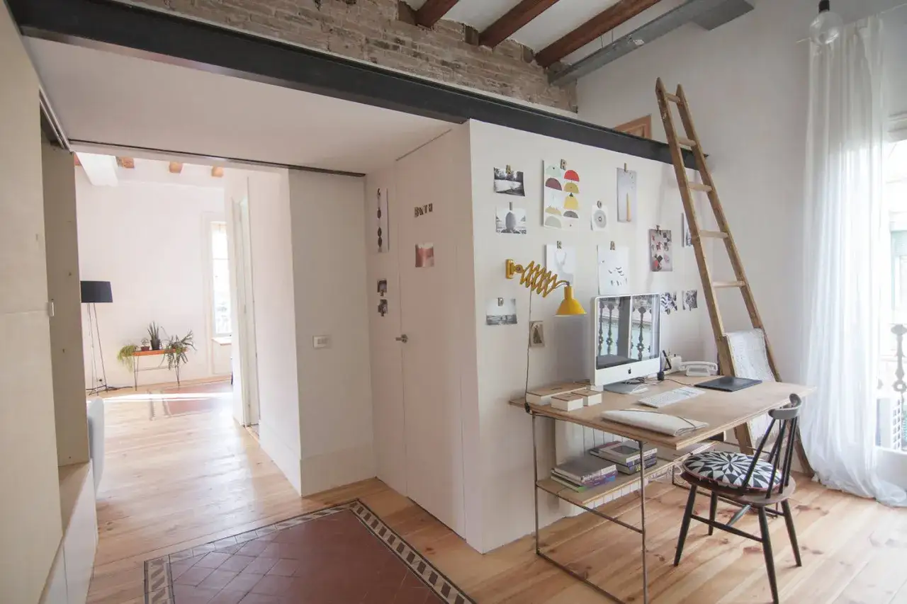 Escritorio con ordenador y una escalera de madera apoyada en la pared, que da acceso a un altillo de madera.