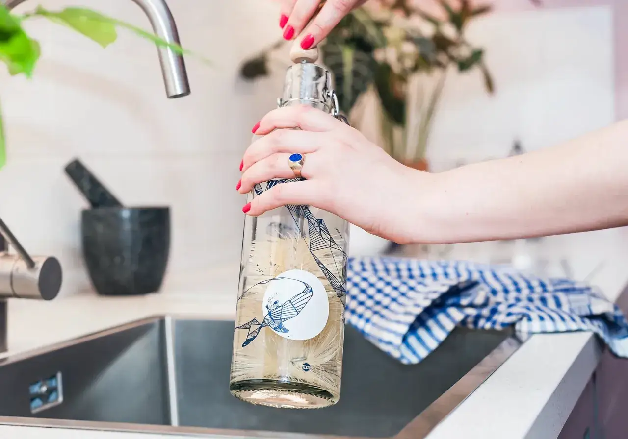 Hände reinigen eine Trinkflasche mit einer Bürste. Ein blauer Ring ziert den Finger.