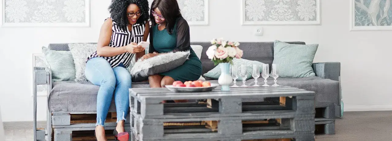 Zwei Frauen lachen &uuml;ber ein Handy auf einem selbstgebauten Palettensofa. Ein Couchtisch aus Paletten steht davor.