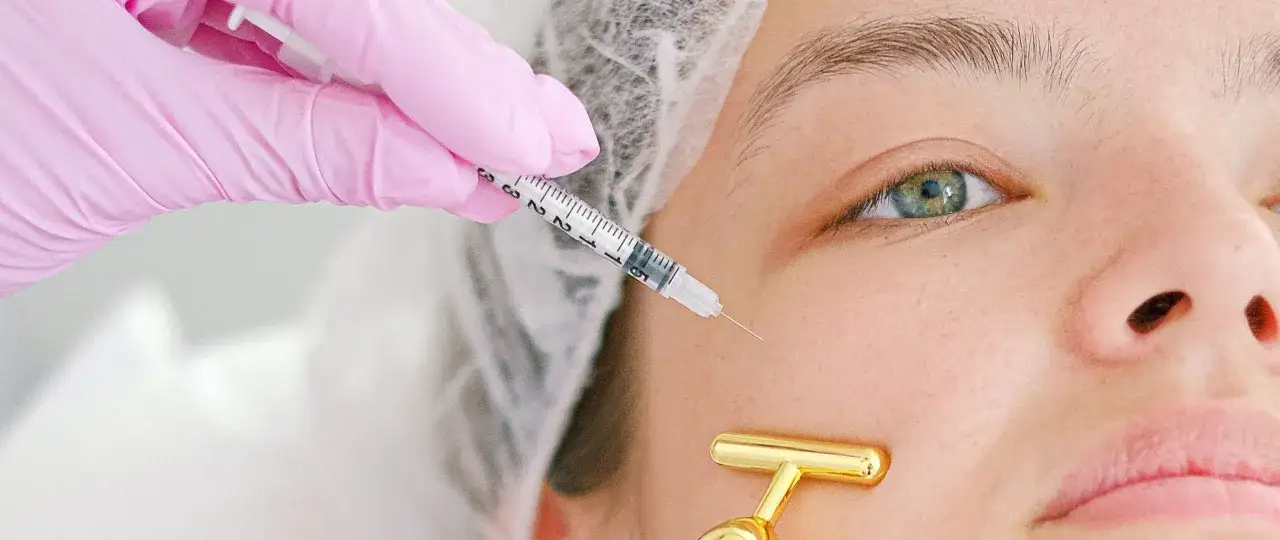 En hand i rosa handske ger en injektion nära ögat. En gyllene massageverktyg ligger på kinden. Botox på recept för en slätare hud.