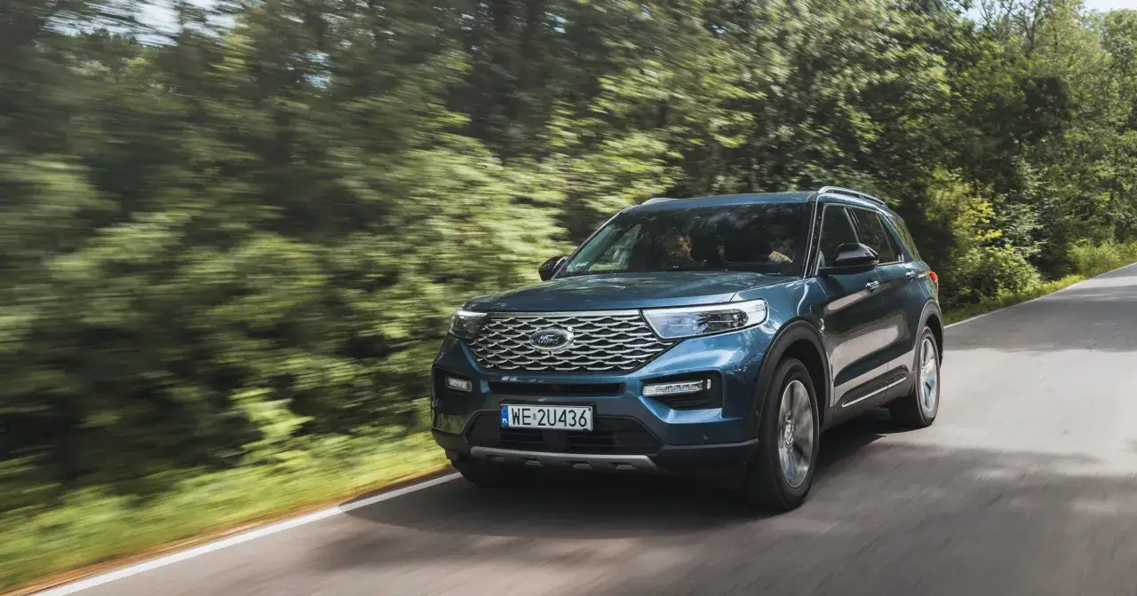 Niebieski ford 7 osobowy pędzi leśną drogą, tworząc wrażenie ruchu dzięki rozmazanym drzewom.