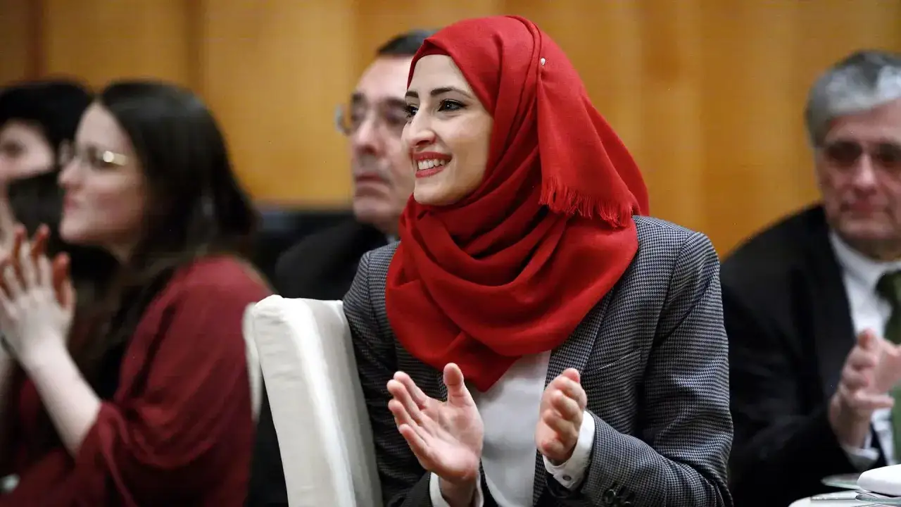 Eine Frau mit einem roten islamischen Kopftuch l&auml;chelt und klatscht. Sie tr&auml;gt einen karierten Blazer.