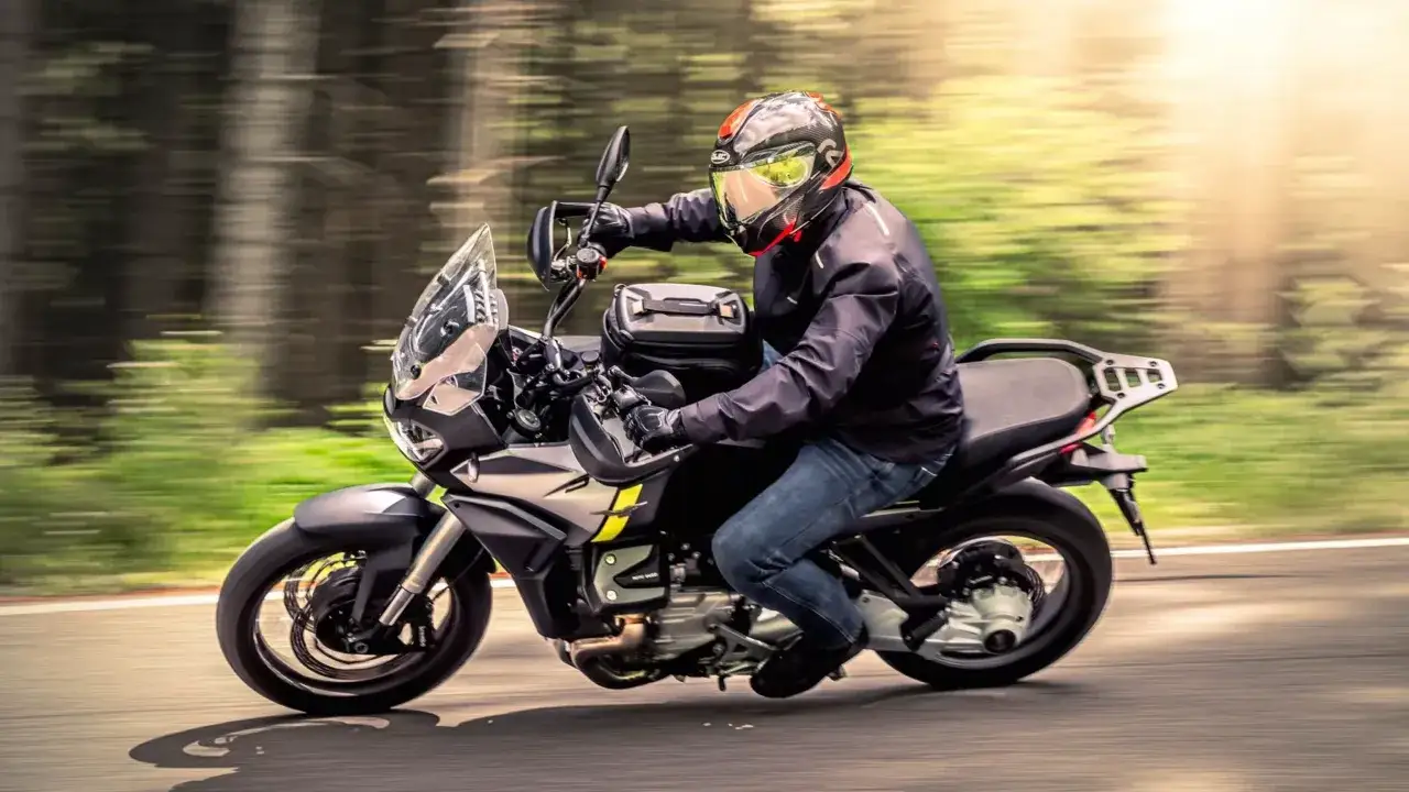 Motocyklista w kasku pędzi na motocyklu Moto Guzzi Stelvio przez leśną drogę, otoczony rozmazaną zielenią i słońcem.