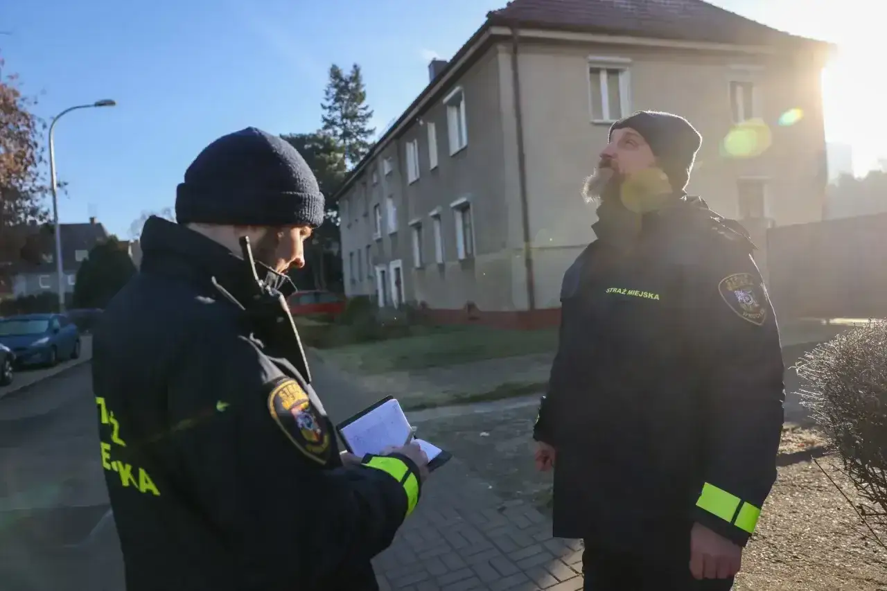 Strażnicy miejscy sprawdzają zgłoszenie o paleniu śmieci. Jeden z nich notuje, drugi obserwuje budynek. Kara za palenie śmieci jest surowa.