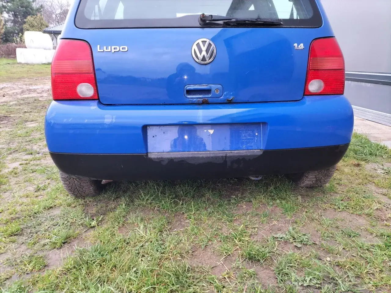 Niebieski Volkswagen Lupo stoi na trawie. Widoczny tył auta z czerwonymi światłami i logo VW.