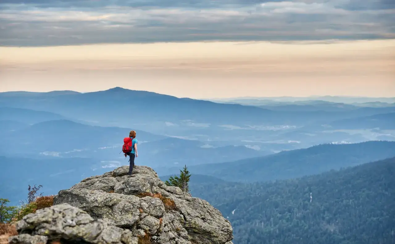 Wanderer auf einem Felsen im Bayerischen Wald, blickt über sanfte Hügel und Wälder. Ein Gefühl von Weite und Ruhe, wie auf einem der 8 Tausender des Bayerischen Waldes.