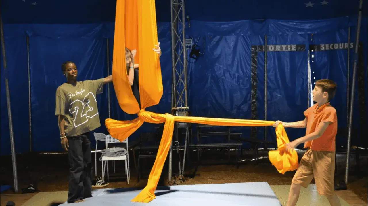 Des enfants apprennent la voltige aérienne lors d'une colonie de vacances artistique, manipulant des tissus orange vifs.