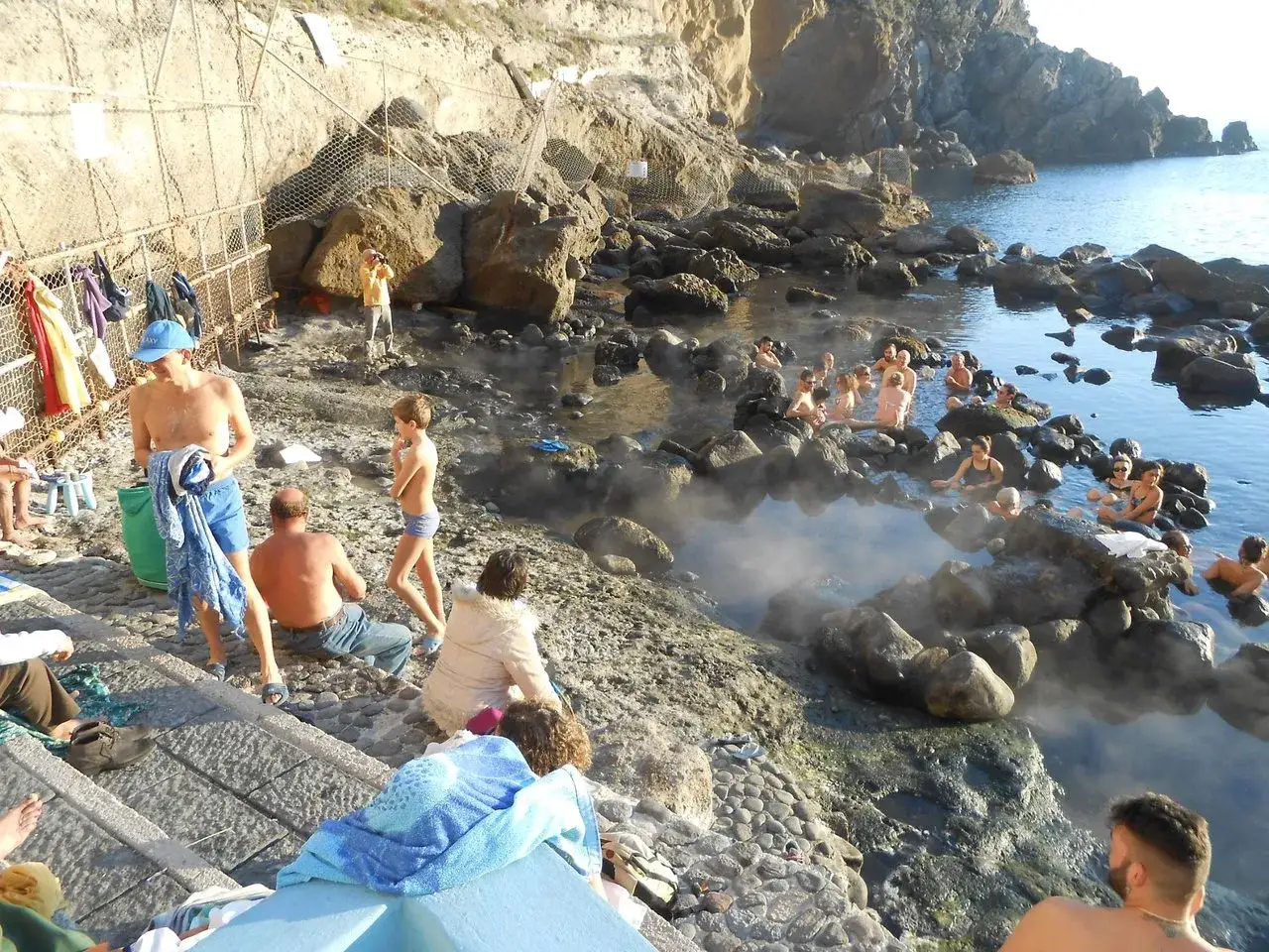 Persone si rilassano nelle acque termali della **baia di Sorgeto**, tra rocce e vapore.