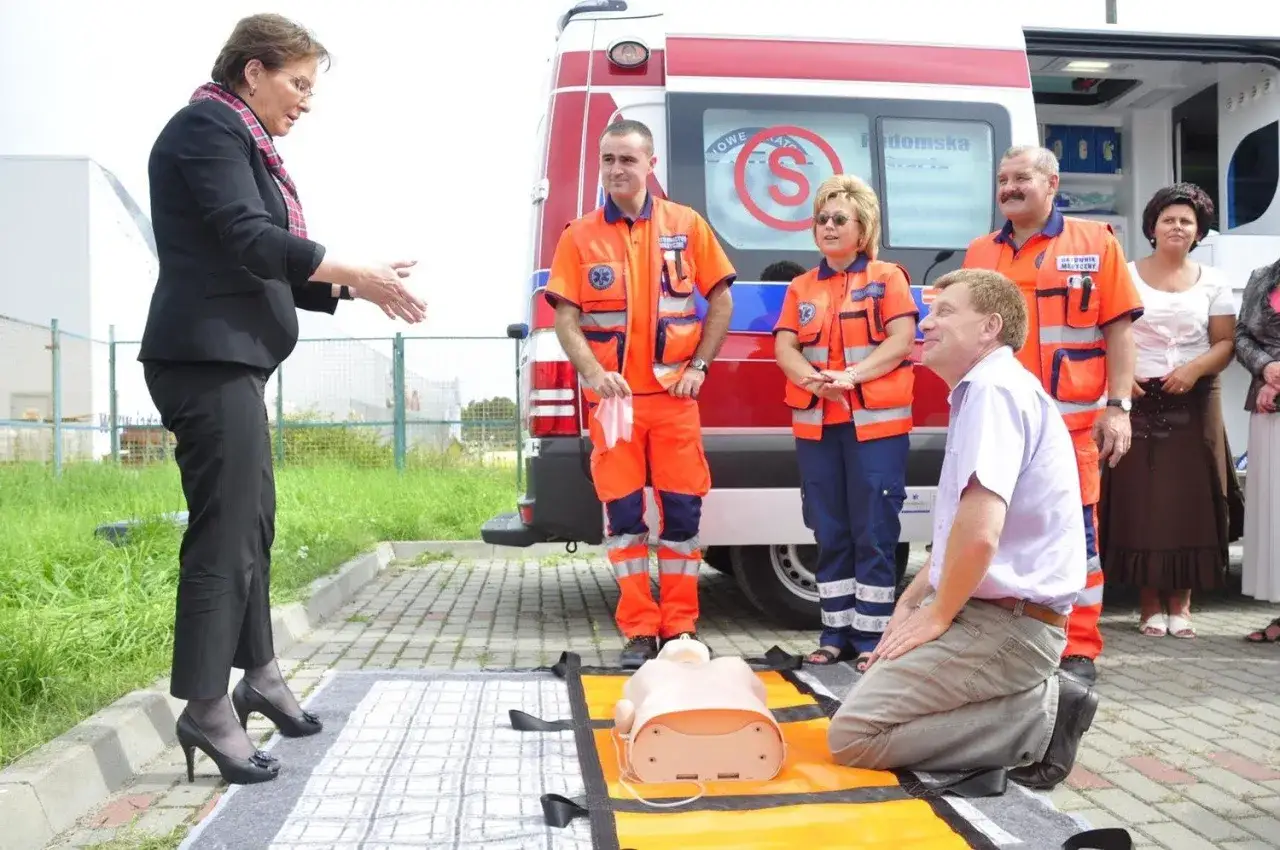 Ewa Kopacz, w otoczeniu ratowników medycznych i karetki, demonstruje resuscytację na fantomie.