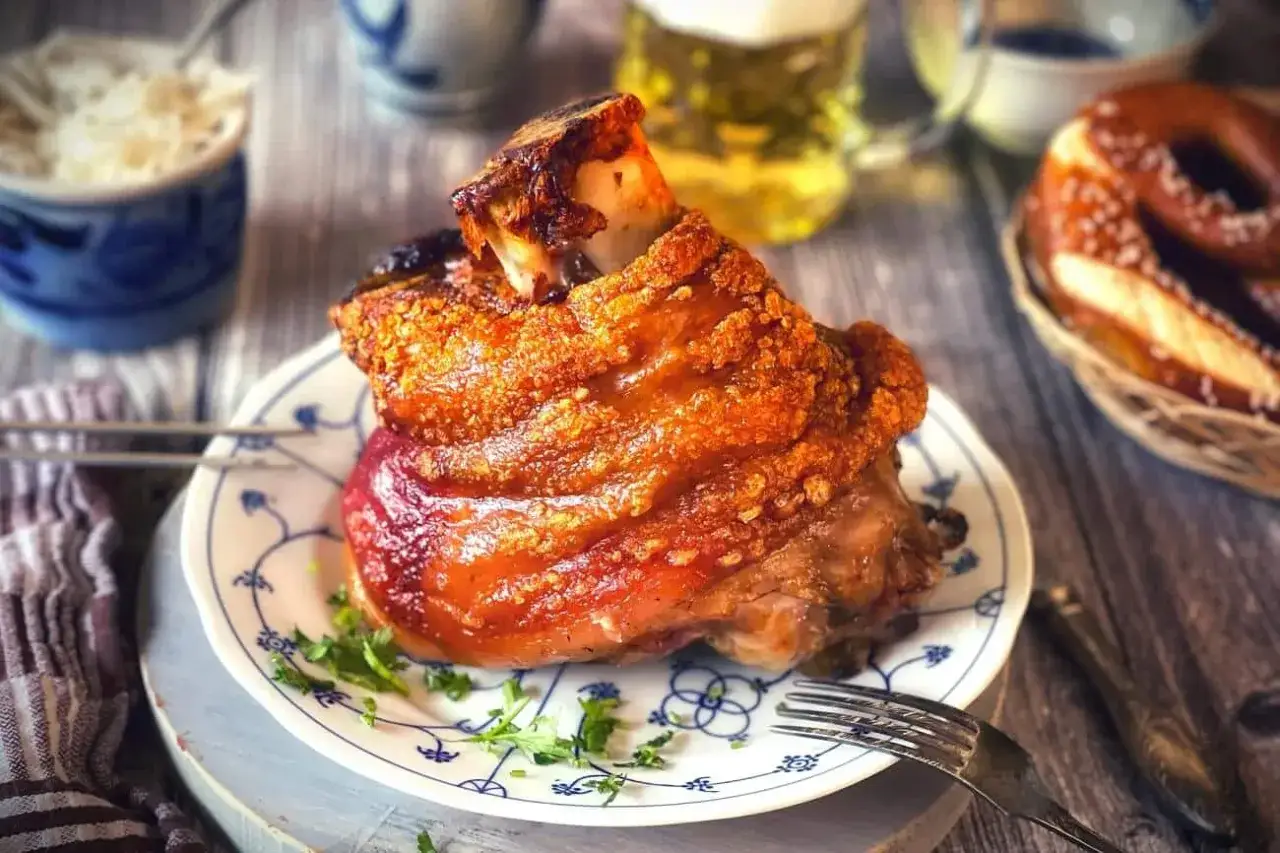 Knuspriger Schweinebraten mit Knochen, ein typisches Oktoberfest Essen, auf einem Teller mit Petersilie. Daneben Bier und Brezel.