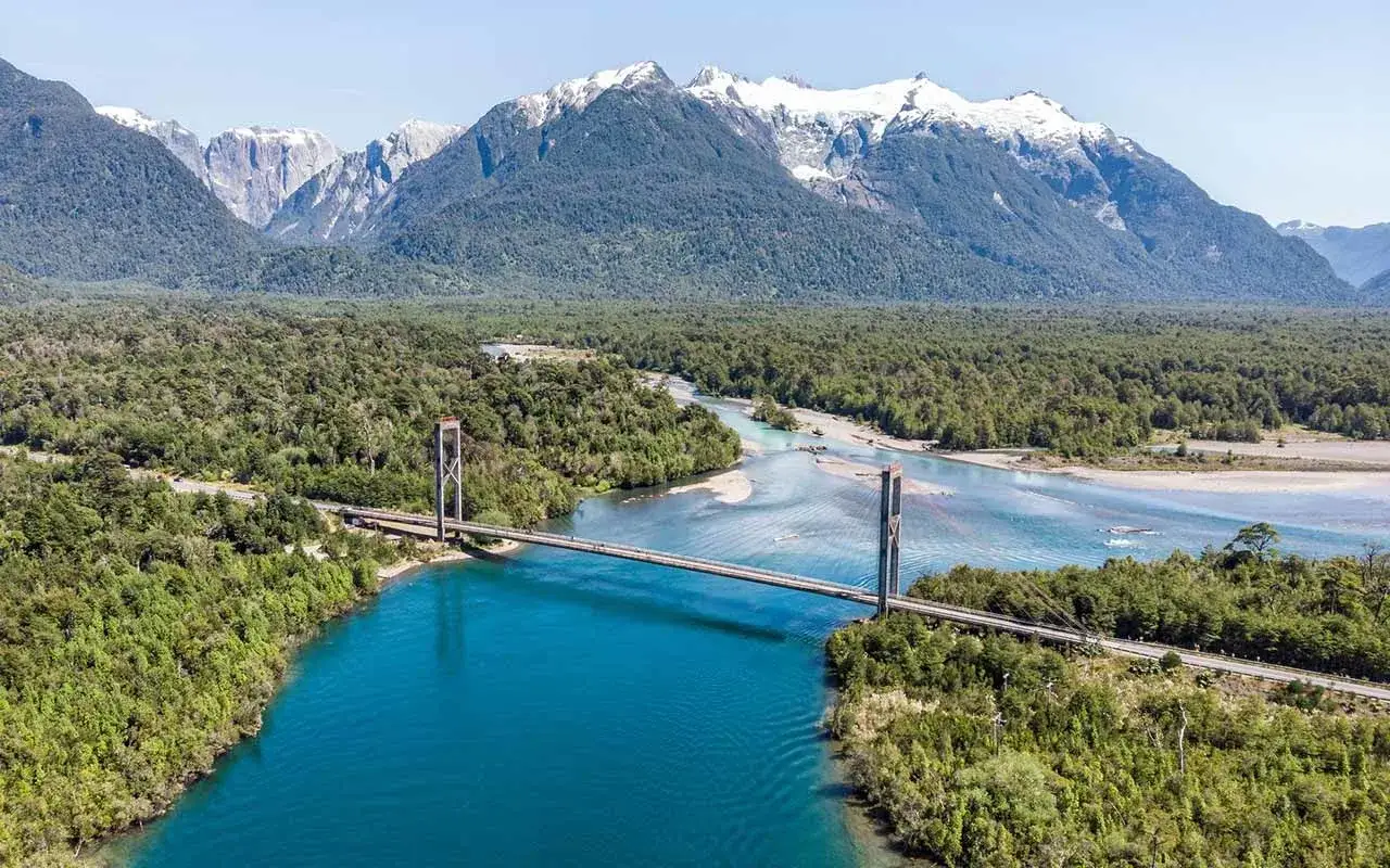 Puente colgante sobre r&iacute;o turquesa, rodeado de bosques frondosos y monta&ntilde;as nevadas. Un tramo ic&oacute;nico de la Carretera Austral.