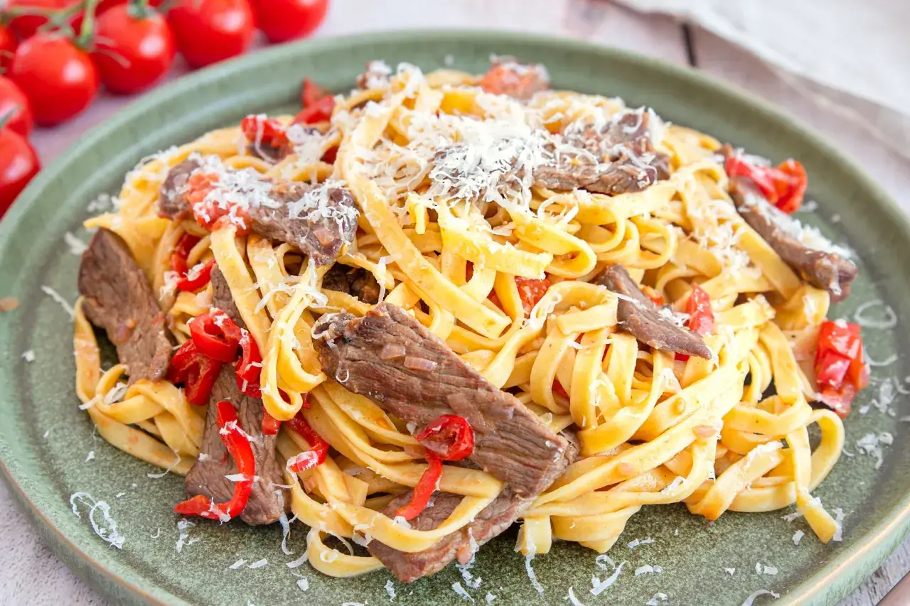 Pasta mit Rinderfiletstreifen, garniert mit geriebenem Parmesan und roten Paprikastücken.