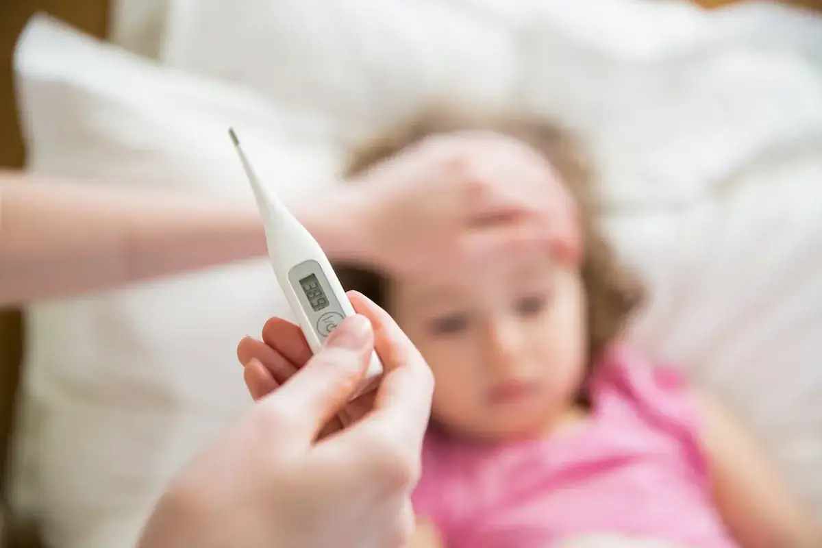 Termómetro muestra 38.9°C, indicando fiebre en niño de 2 a 3 años. Una mano sostiene el termómetro cerca de la frente del niño.