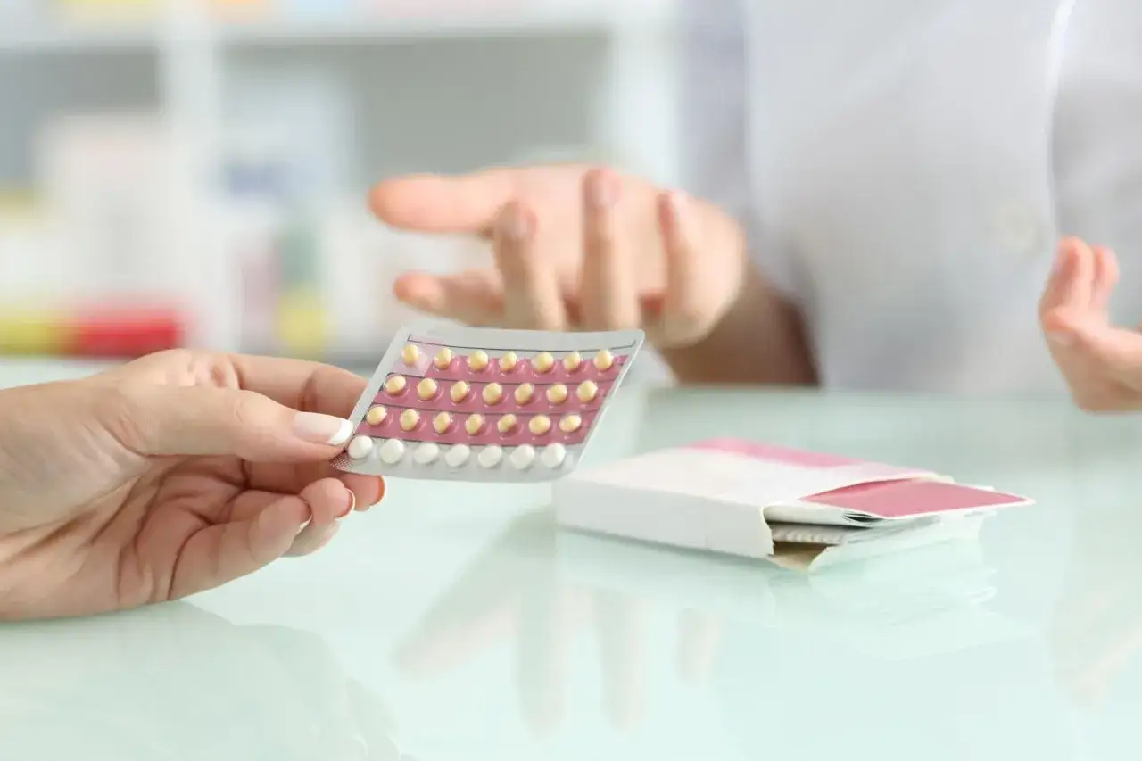 Dłoń trzyma opakowanie tabletek antykoncepcyjnych, podobnych do Qlaira. Farmaceuta wyjaśnia coś w tle.