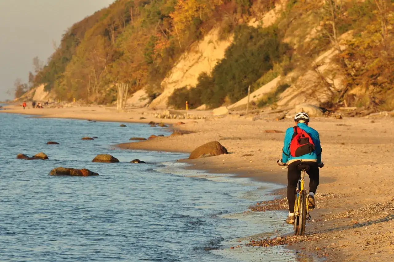 Rowerzysta przemierza piaszczyste plaże zachodniego wybrzeża Bałtyku. W tle widać klif porośnięty jesiennymi drzewami.