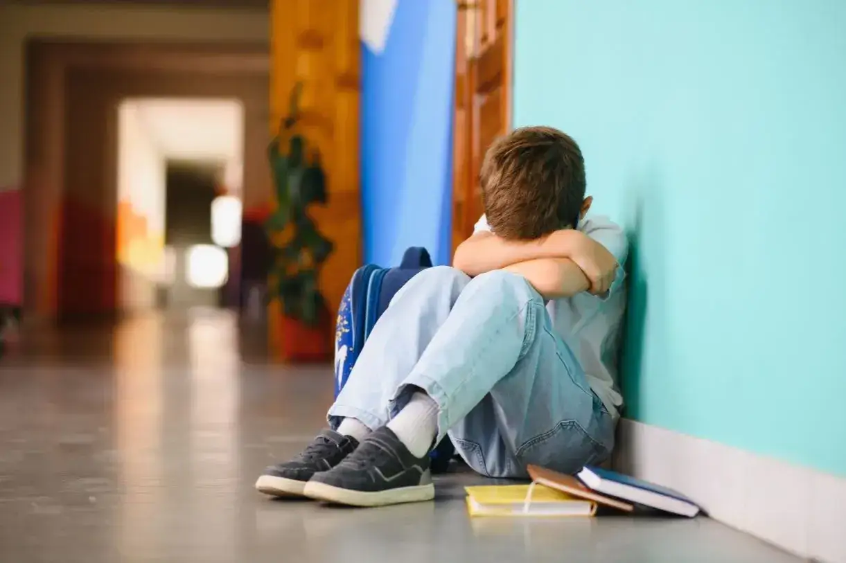 Chłopiec siedzi skulony w kącie korytarza, przytulając plecak. Wygląda na to, że doświadcza **bullyingu**.