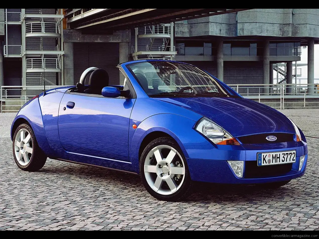 Niebieski Ford Streetka z otwartym dachem stoi na brukowanym placu przed nowoczesnym budynkiem.