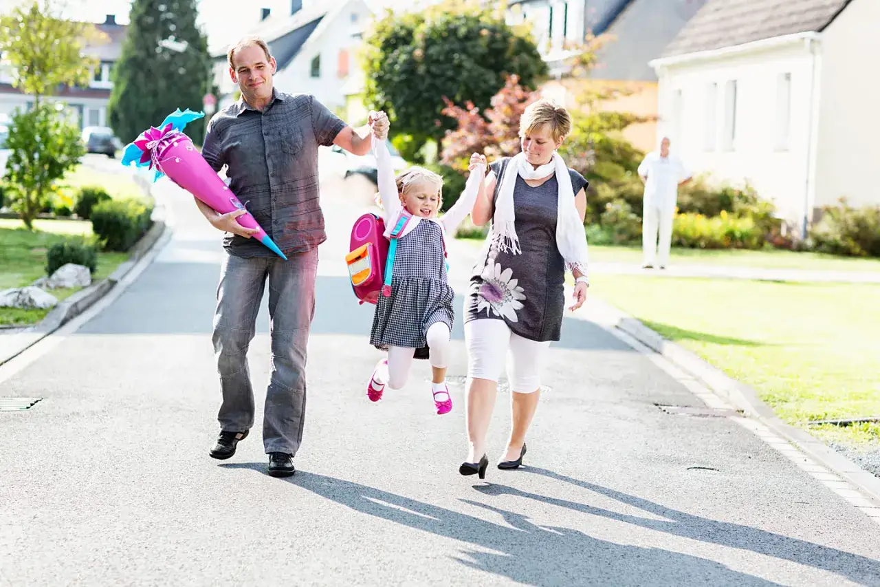 Familie geht zur Einschulung. Vater trägt Zuckertüte, Mutter und Kind sind fröhlich. Das Kind trägt ein schickes Kleid, perfekt, was zur Einschulung anziehen.