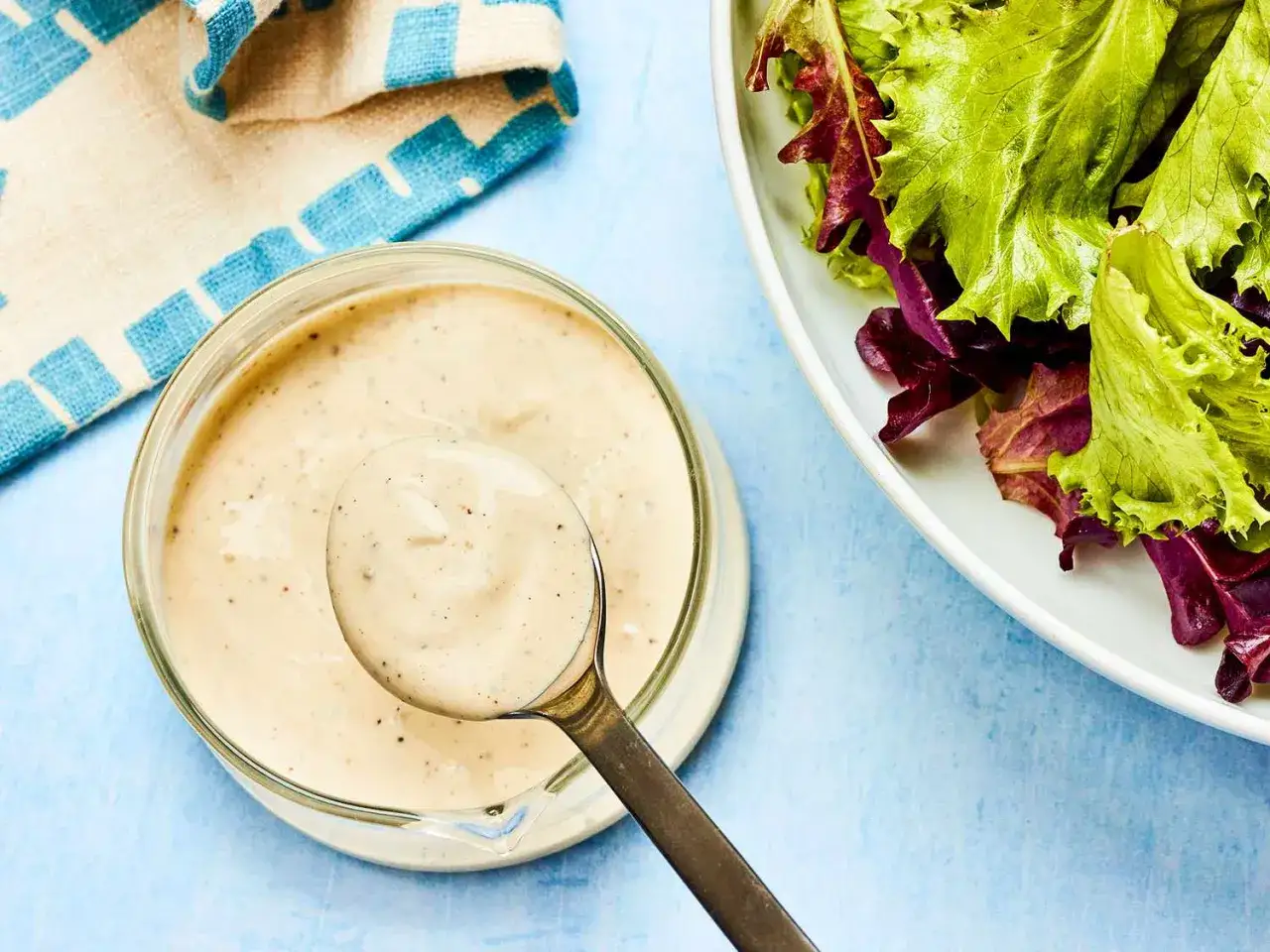 En sked krämig dressing till sallad lyfts ur en skål. Bredvid syns en fräsch sallad med gröna och röda blad.