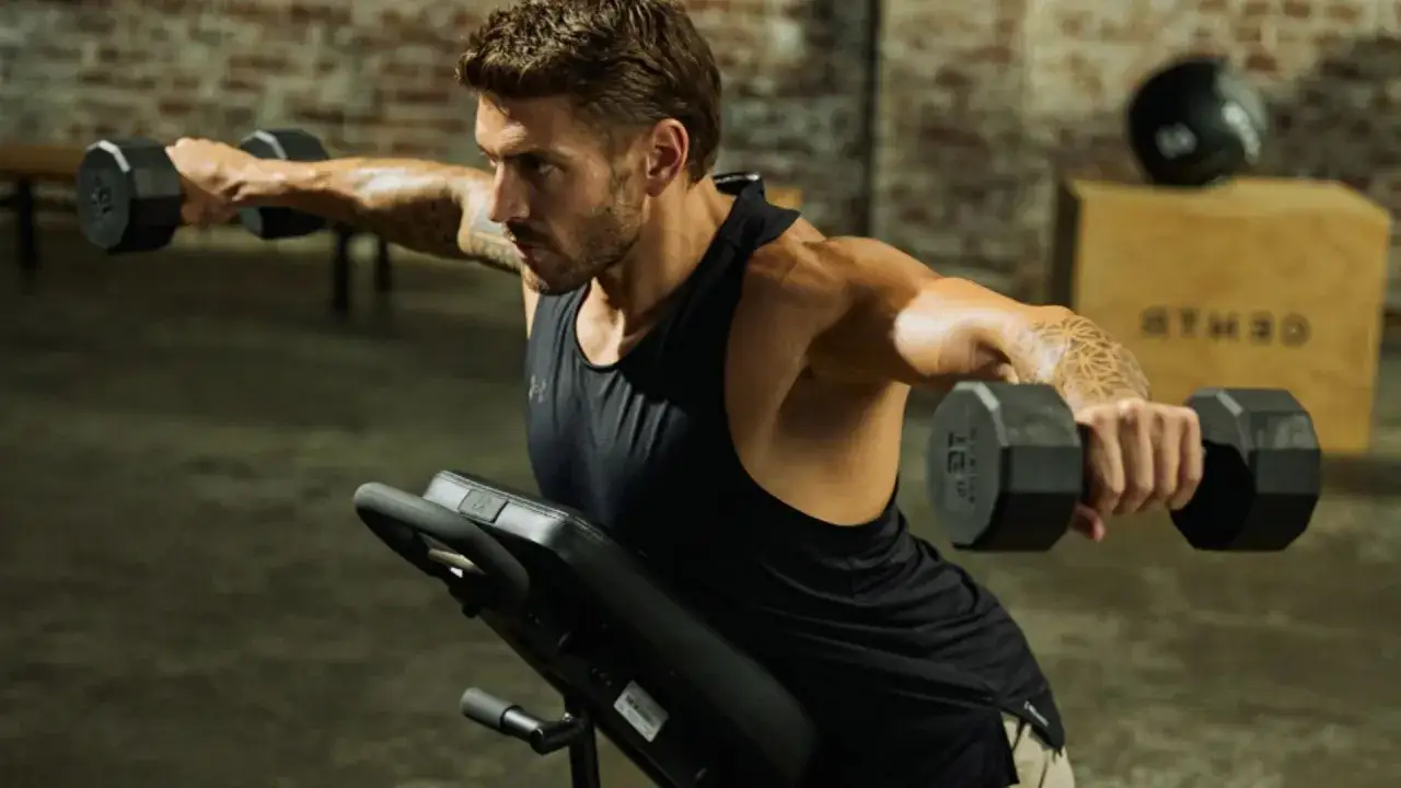 Uomo concentrato mentre solleva pesi con manubri, allenamento spalle in palestra.