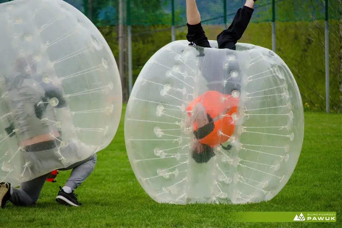Gracze w kulach do bubble football zderzają się na trawiastym boisku. Jedna osoba jest do góry nogami w swojej bańce.