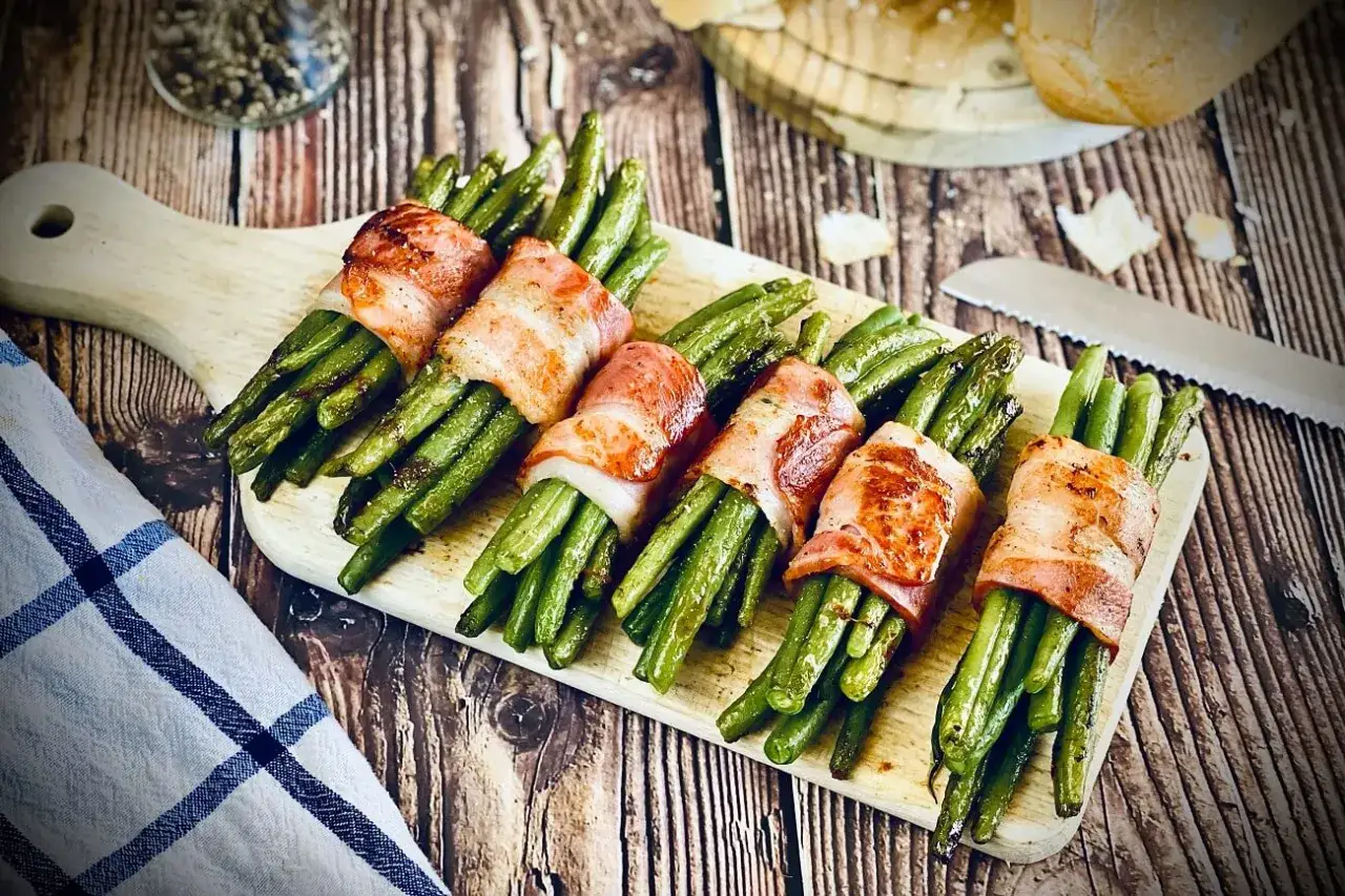 Sechs Bündel grüner Bohnen, umwickelt mit knusprigem Speck, liegen auf einem Holzbrett. Köstliche Bohnen im Speckmantel, bereit zum Genuss.
