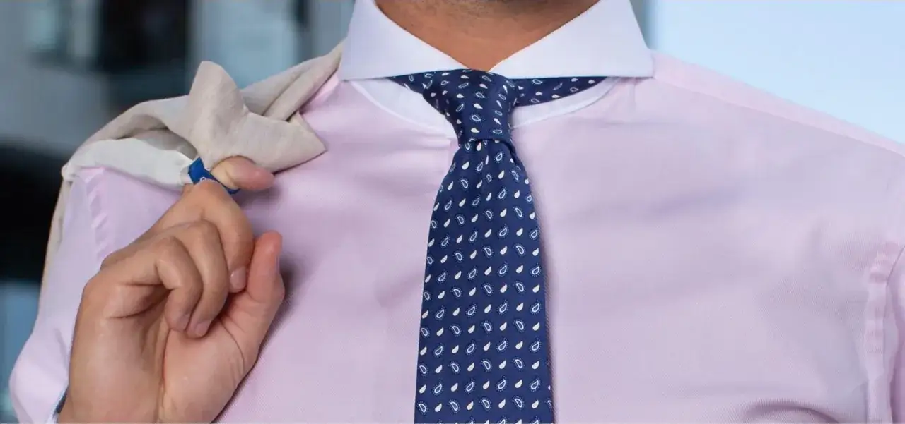 En man med ljusrosa skjorta och mörkblå slips med paisley-mönster. En kavaj ligger över axeln. Olika kragar namn kan vara svårt att välja, men denna skjorta har en klassisk krage.