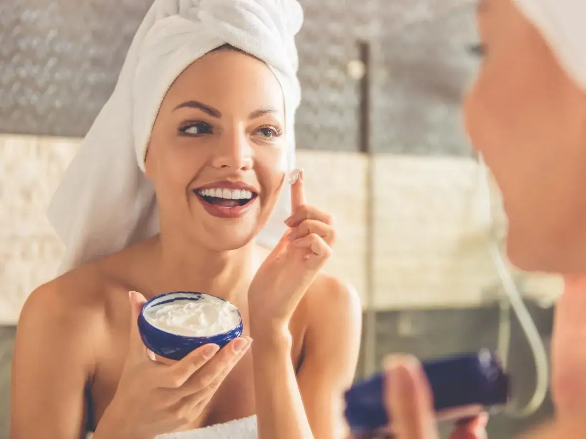 Donna sorridente con asciugamano in testa applica crema sul viso, contrastando la desquamazione della pelle.