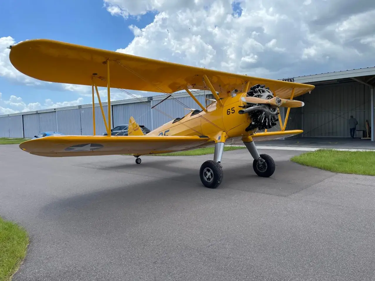 Ein gelbes Stearman Flugzeug mit der Nummer 65 steht auf einem Rollfeld vor einer Flugzeughalle.