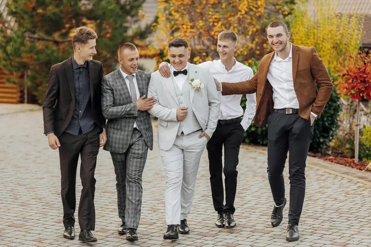 Pięciu mężczyzn w eleganckich garniturach idzie razem. Idealna inspiracja, jak ubrać się na ślub, od klasycznej szarości po brązową marynarkę.