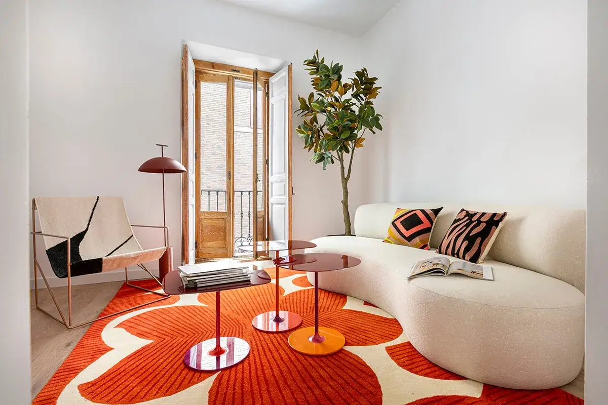Sof&aacute; curvo blanco, alfombra naranja con flores, sill&oacute;n moderno y mesa auxiliar, combinando diversos estilos de dise&ntilde;o.
