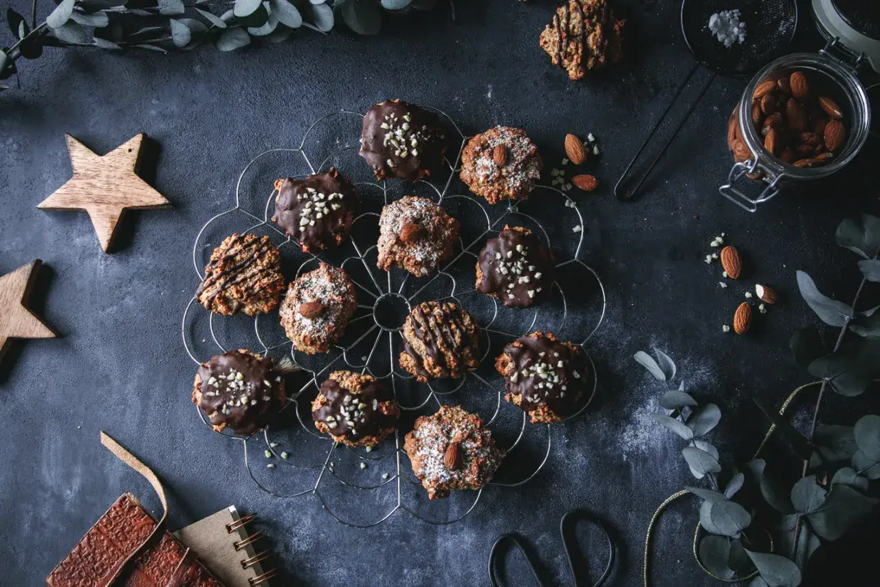 Schokoladen- und mandelverzierte Kekse auf einem Gitter, inspiriert von alten Lebkuchen Rezepten. Daneben Mandeln, ein Glas, Sterne und Zweige.