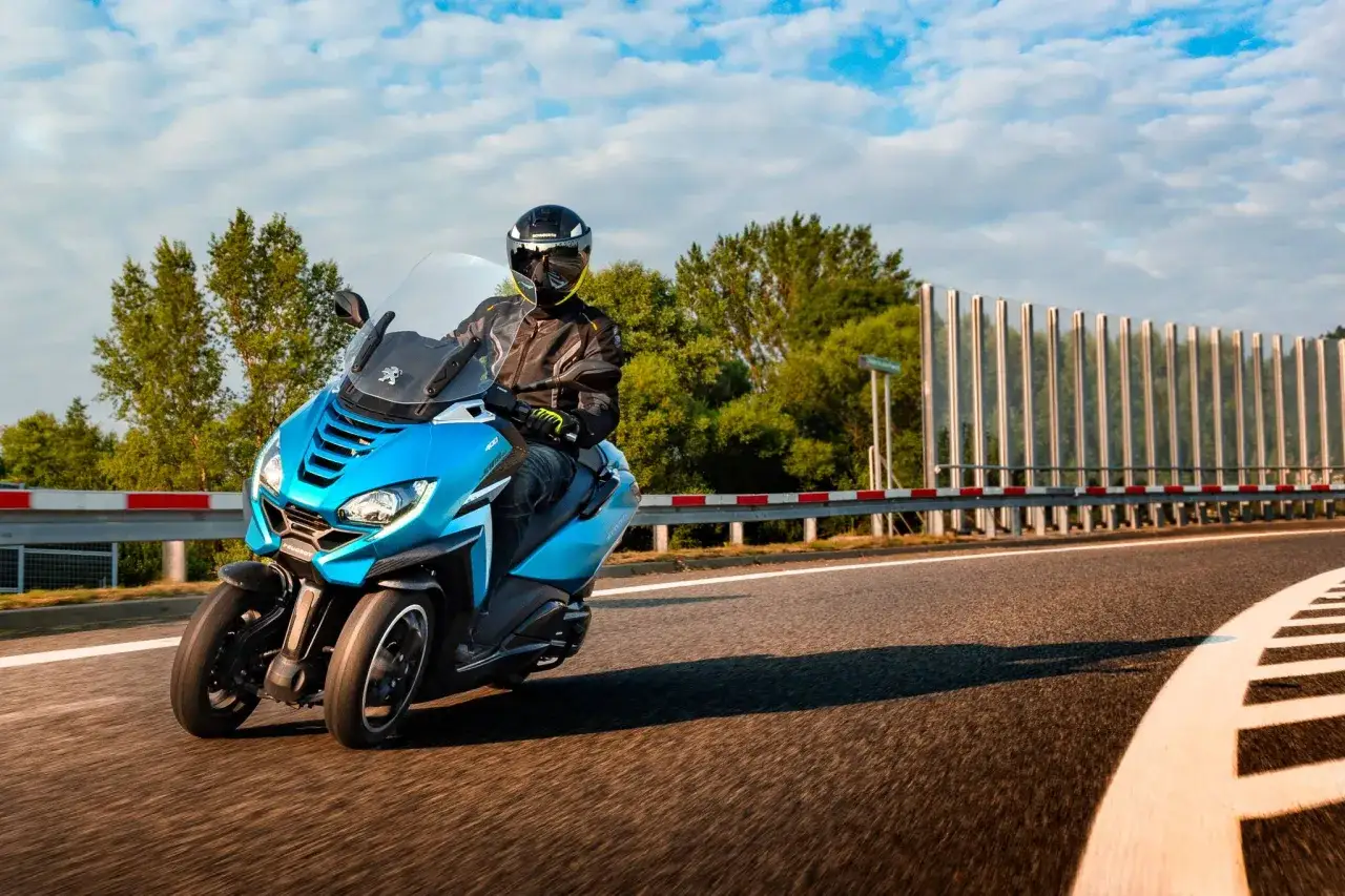 Niebieski Peugeot Metropolis 400 z kierowcą w kasku pędzi po zakręcie drogi, otoczony zielenią i błękitnym niebem.