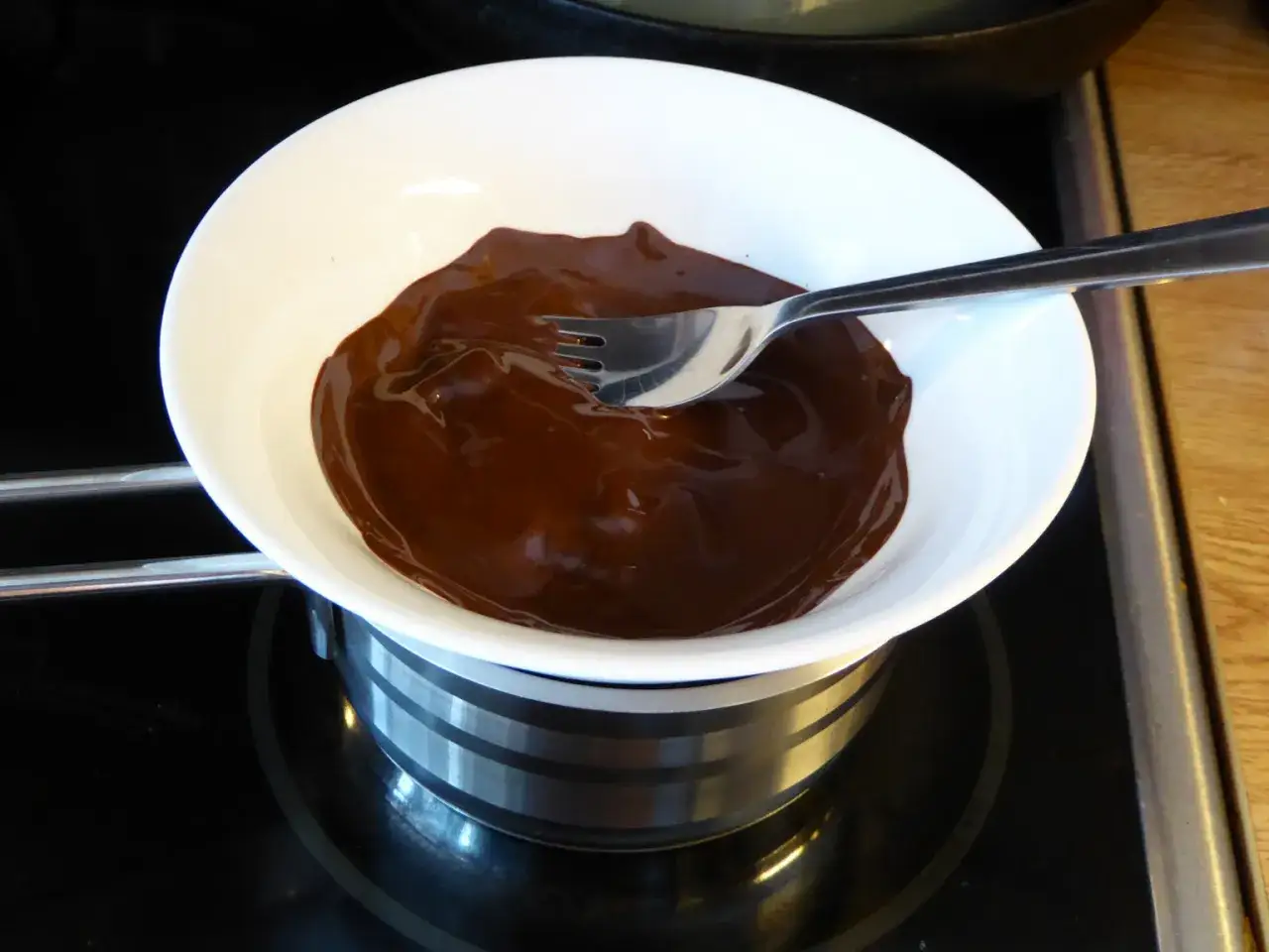 Sm&auml;lt choklad i ett vattenbad. En gaffel r&ouml;r om den glansiga, m&ouml;rka chokladen i en vit sk&aring;l.
