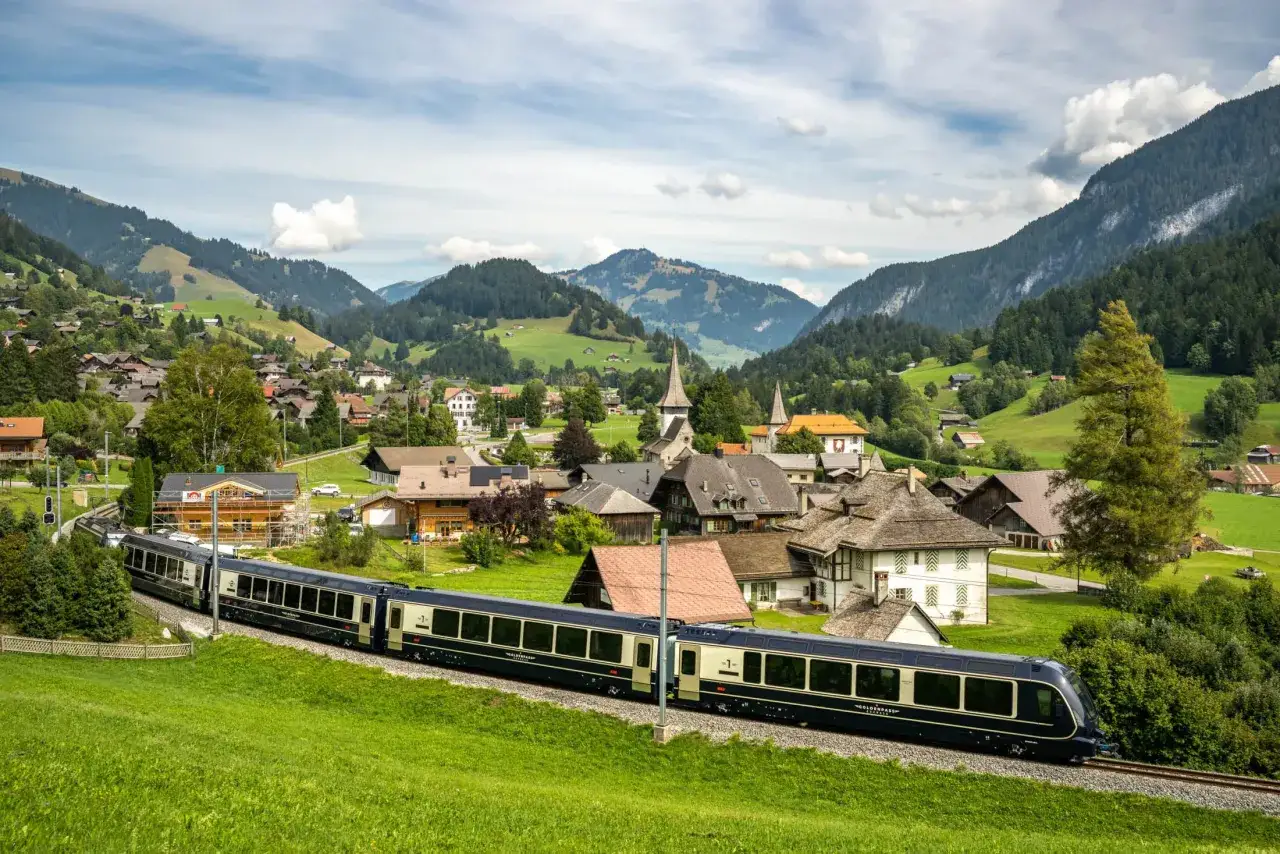 A train travels through a Swiss village on the Golden Pass Montreux to Interlaken route, with green hills and mountains in the background.