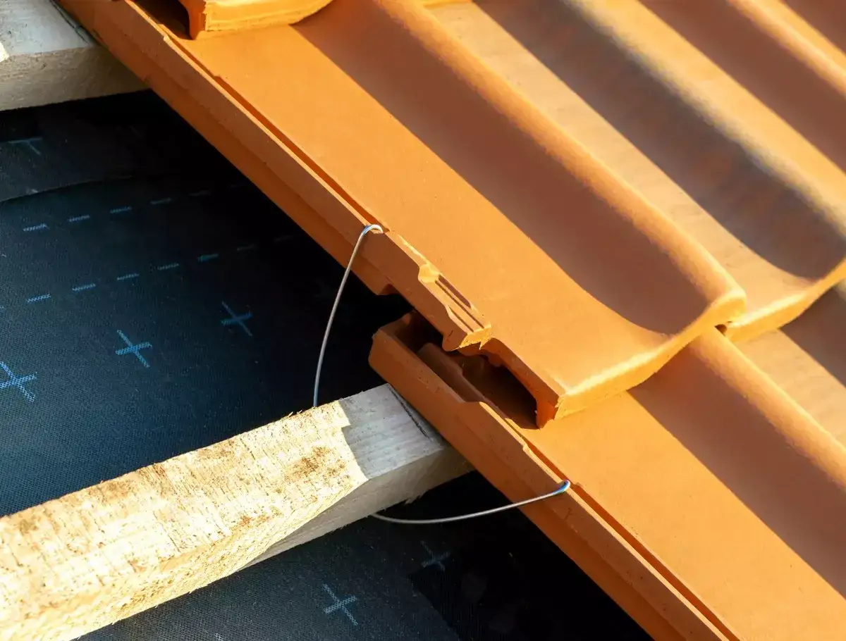 Zbliżenie na dachówkę ceramiczną, gdzie widać drutowe spinki do dachówki mocujące ją do drewnianej łaty. Pod spodem czarna membrana dachowa.