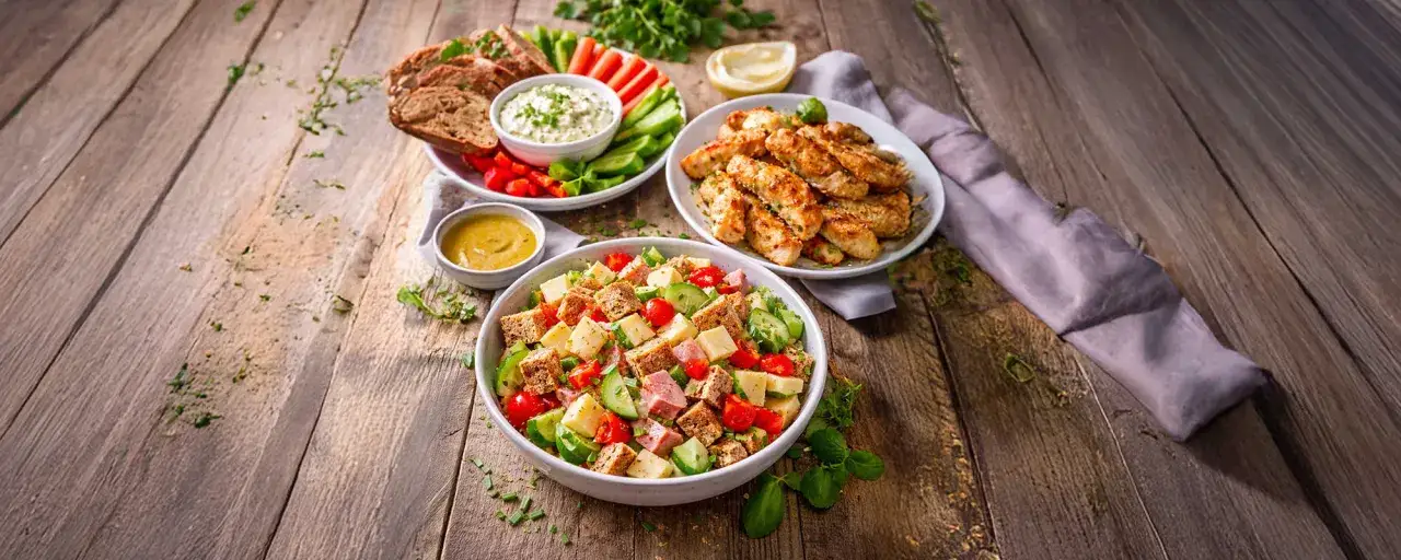 Eine **gesunde Brotzeit** mit einem bunten Salat, gegrillten Hähnchenstreifen, Brot mit Dip und Gemüsesticks.