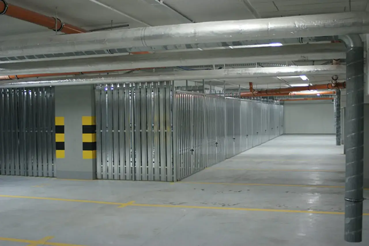 Rząd metalowych komórek lokatorskich w podziemnym garażu. Widać rury instalacyjne na suficie i żółte linie na posadzce.
